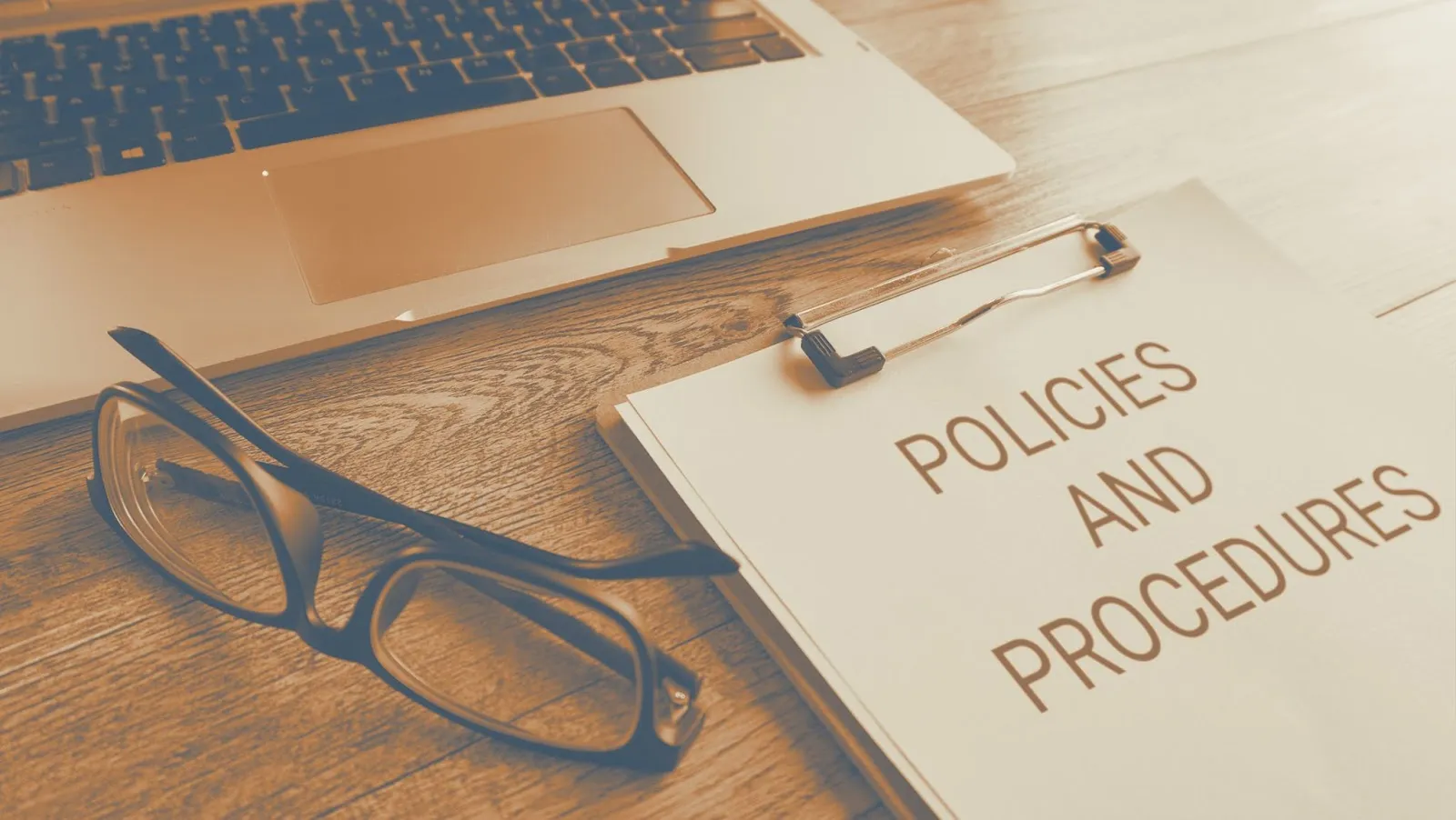 Clipboard labeled Policies and Procedures on a wooden desk next to glasses and a laptop