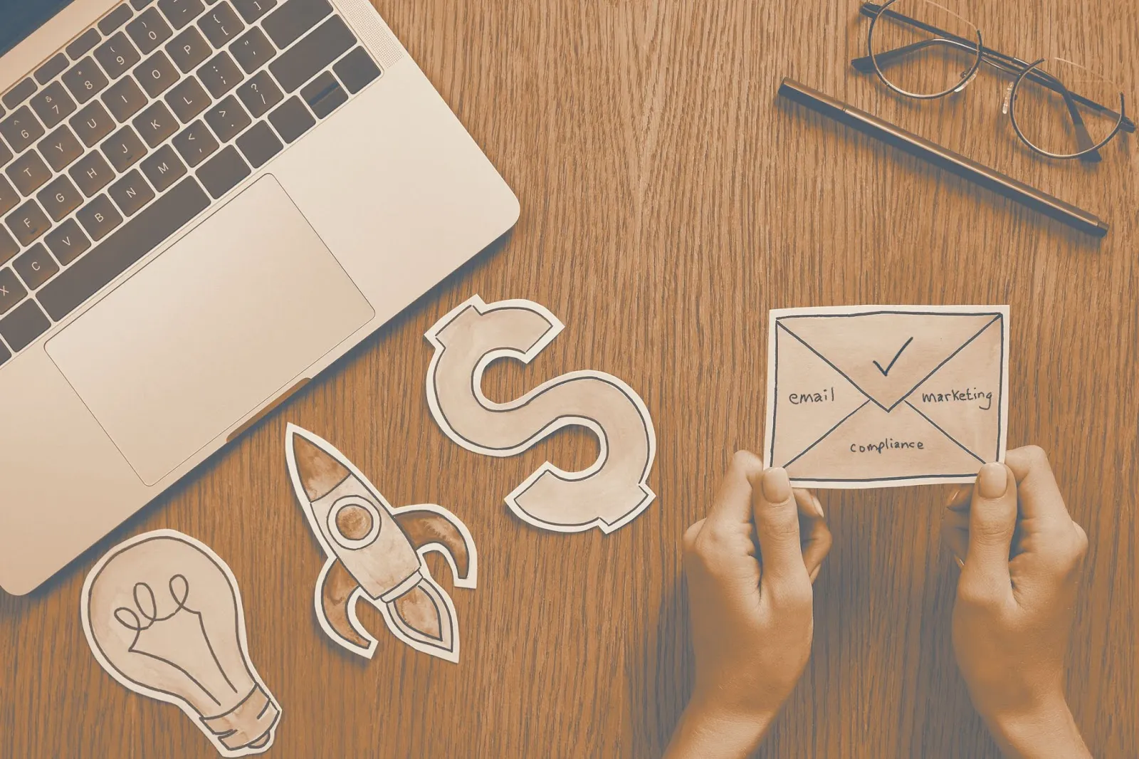 Flat lay desk with laptop, paper cutouts of a lightbulb, rocket, dollar sign, and email compliance envelope