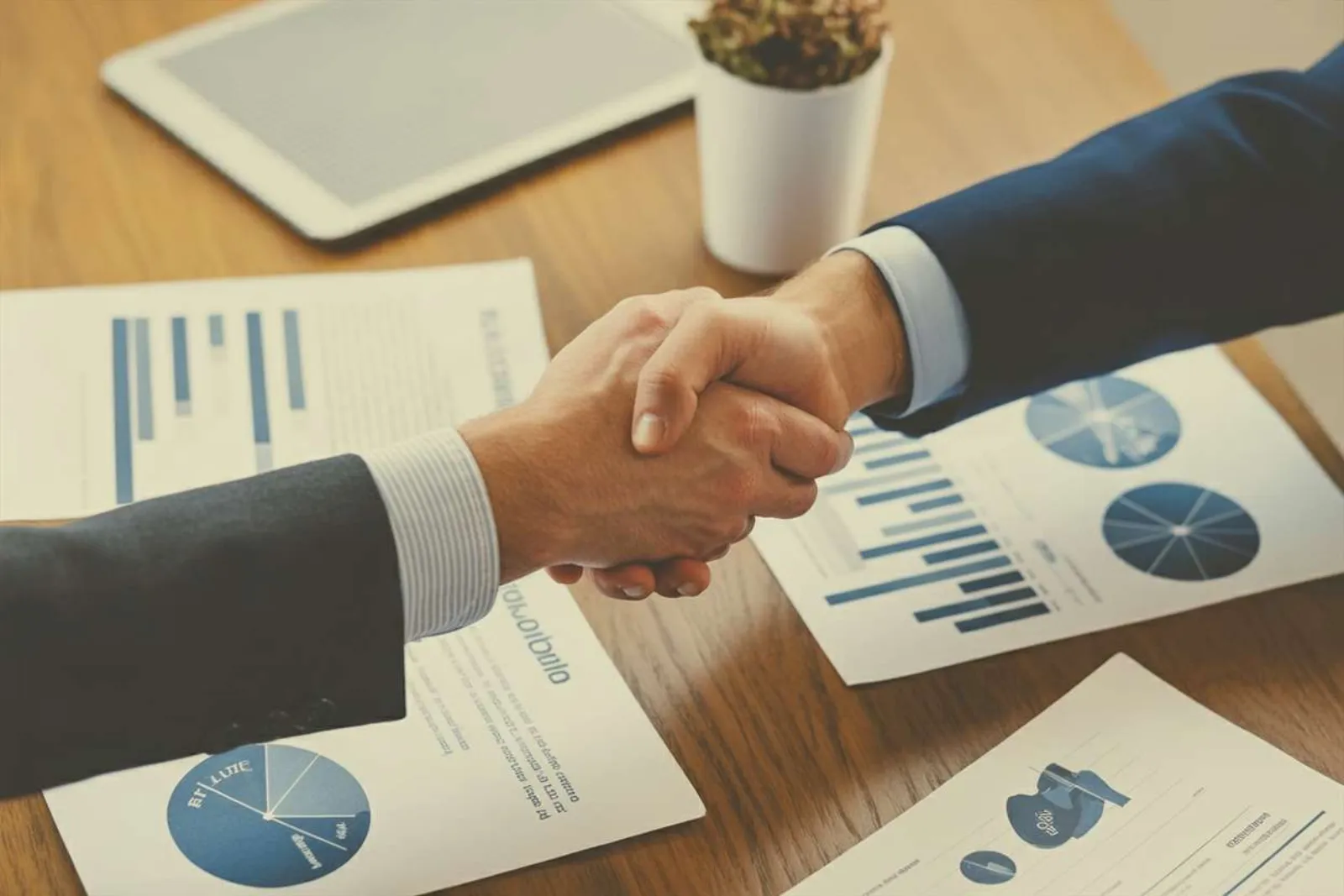 Two professionals shaking hands over financial reports with bar and pie charts